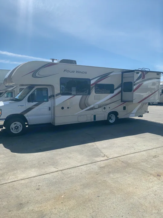 This image features a beige and white Four Winds Class C RV rental with its slide-out extended, parked on a concrete lot.. Thor Motor Coach Four Winds 2020