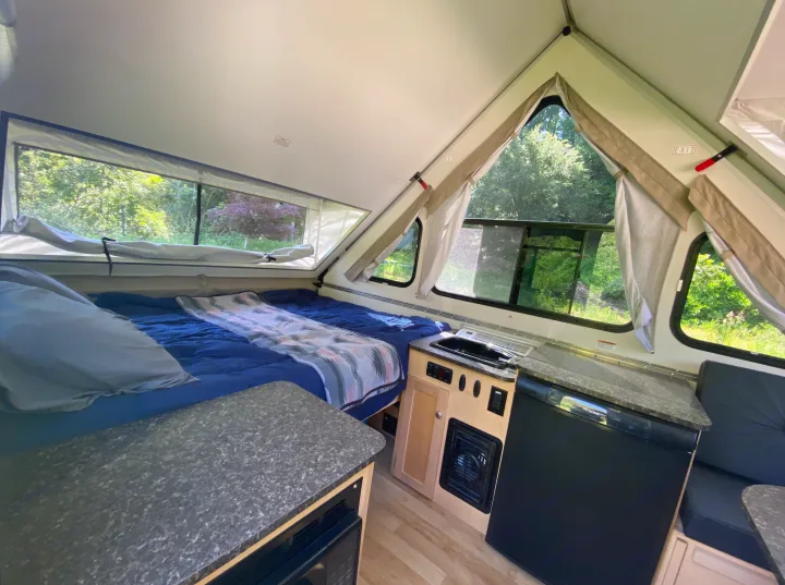 A-frame RV rental interior: A cozy bed, compact kitchenette with fridge & microwave, and bright windows overlook nature.. A-Liner Classic 2019