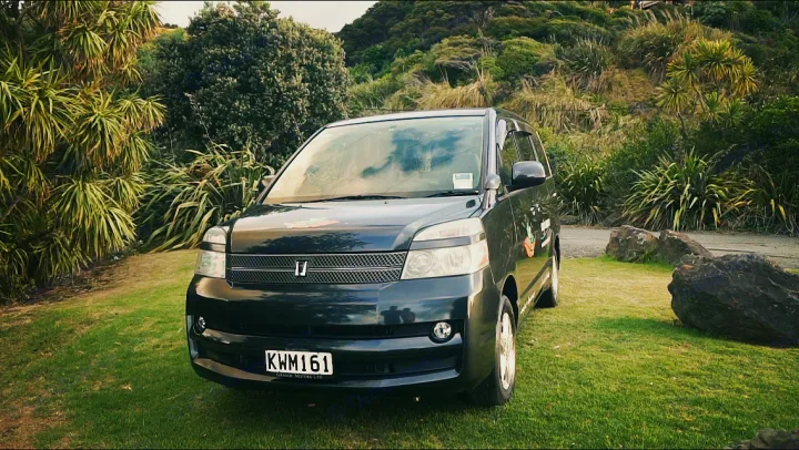 Dark grey minivan (KWM161) parked on grass. This compact RV rental is set against lush New Zealand hills.. Toyota Voxy 2006