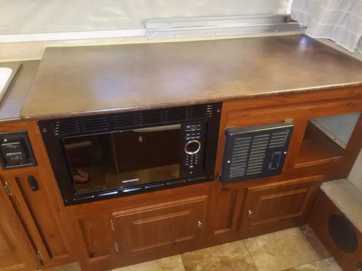 This RV rental interior shows a countertop, a Greystone microwave, and wooden cabinets, with a Suburban furnace vent on the right.. Forest River Rockwood Premier 2013