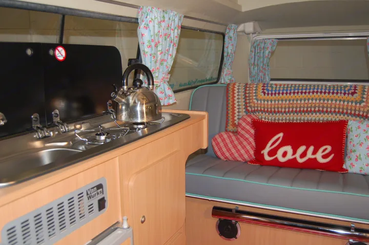Compact RV rental interior: a gleaming kitchenette, cozy bench seat with "love" pillow, and cheerful floral curtains.. Volkswagen 2.0L T2 Late Bay 1975