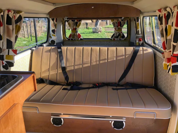 Retro camper van rv rental interior: tan bench seat with seatbelts, bright curtains, view outside, and built-in speakers.. Volkswagen 2.0L T2 Late Bay 1975