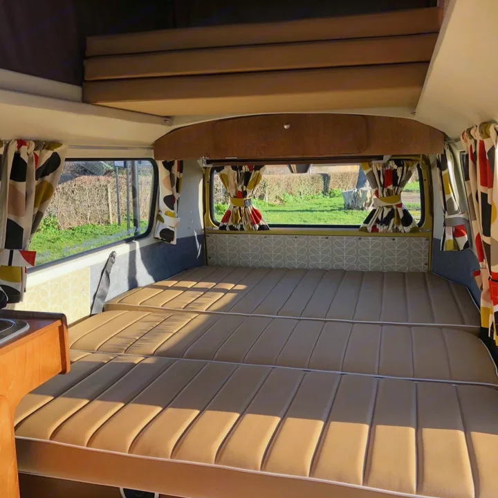 Experience this RV rental: a large tan bed, colorful patterned curtains, and an overhead sleeping area in a bright camper.. Volkswagen 2.0L T2 Late Bay 1975