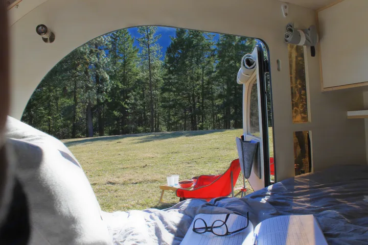 Inside an rv rental: a comfy bed and open door frame a sunny forest meadow with an outdoor table & chair setup.. DROPLET 58 2019