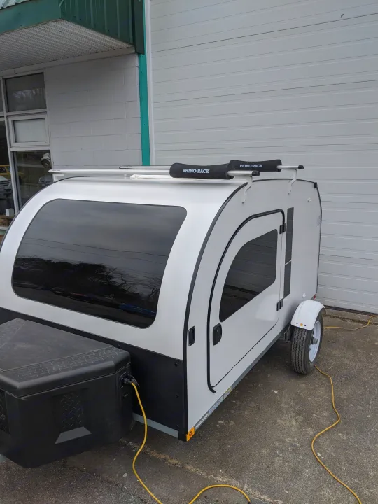 This white teardrop travel trailer, an RV rental with a Rhino-Rack, is parked on asphalt. A compact camper perfect for road trips.. DROPLET 58 2019