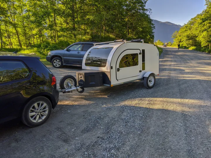 A white teardrop **rv rental** is connected to a car on a scenic gravel road, surrounded by lush trees and mountains for an outdoor adventure.. DROPLET 58 2019