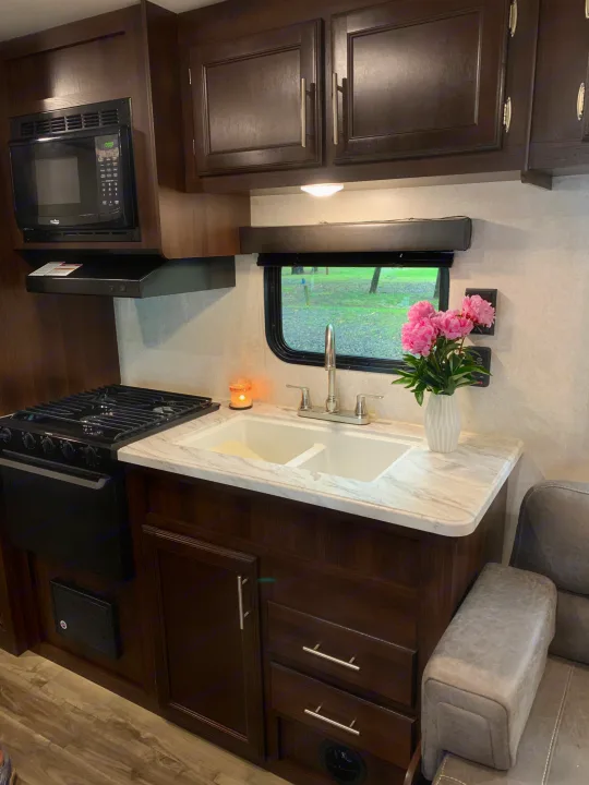 RV rental kitchen: Dark wood cabinetry, microwave, stovetop, double sink with pink flowers by the window, and cozy gray seating.. Jayco Jay Flight 2019