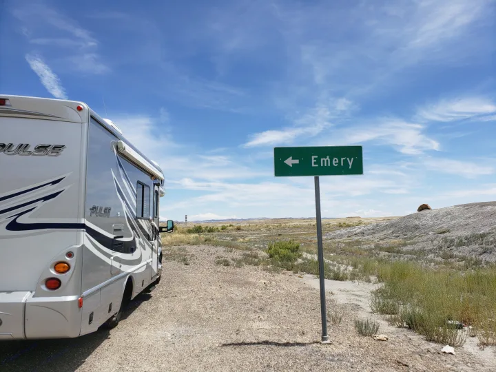 An RV rental is parked on a dirt road by a green sign pointing to Emery, with a desert landscape under a blue sky.. Fleetwood Pulse 2009