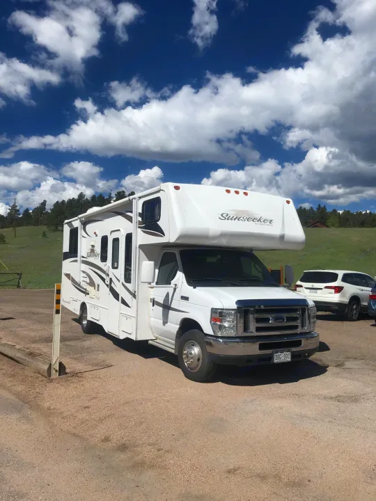 A white Sunseeker RV rental parked on a gravel lot, with green hills and a blue sky with white clouds.. ForestRiver Sunseeker 2012