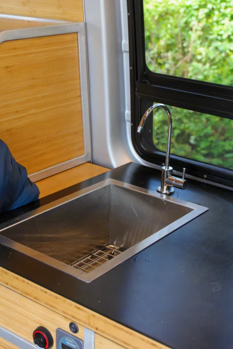 Inside an RV rental, a compact kitchen with a stainless steel sink, black countertop, bamboo cabinetry, and a window with screen.. Mercedes-Benz Sprinter 2018