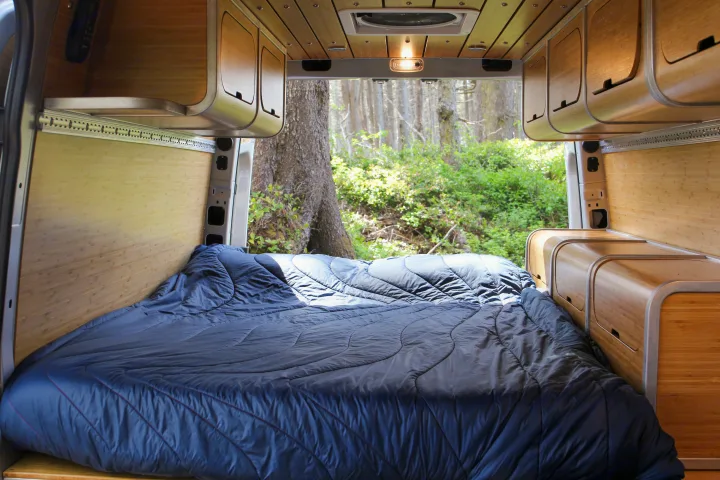 This RV rental offers a cozy, well-appointed interior with a large bed, bamboo accents, and a picturesque forest backdrop.. Mercedes-Benz Sprinter 2018