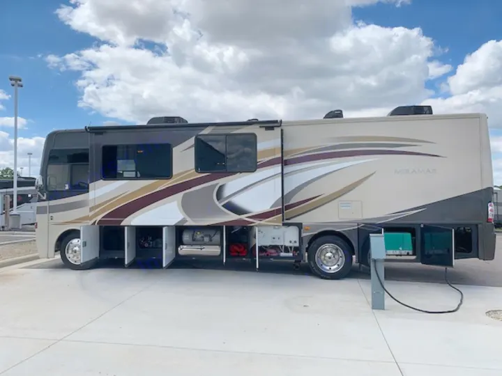 A large beige Miramar RV rental is parked with storage bays open, connected to power, under a cloudy sky.. Thor Motor Coach Miramar 2014