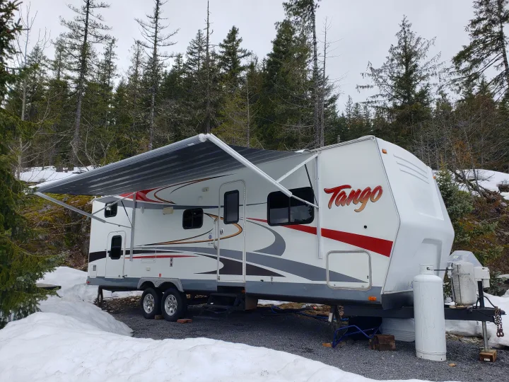 A white Tango travel trailer RV rental parked in a snowy forest campsite with its awning extended, ready for winter camping.. Pacific Coachworks Tango 2010
