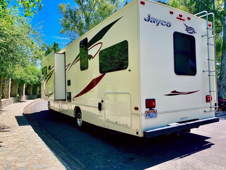 A cream-colored Jayco RV rental is parked with extended slide-outs on a sunny road by a stone path.. Jayco Redhawk 2016