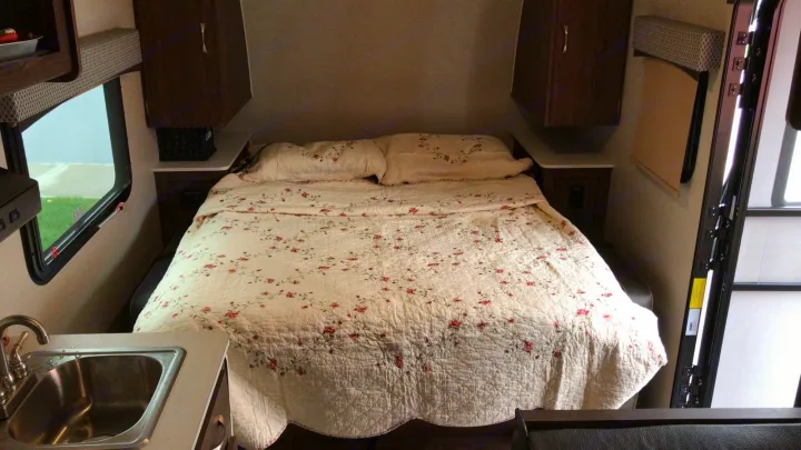 Cozy RV rental interior features a floral bed, overhead storage, window views, and a compact kitchen sink area.. Forest River Rockwood 2018