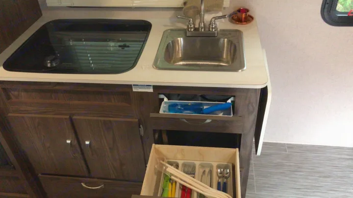 RV rental kitchen: a compact space with a sink, covered stovetop, and open wooden drawer revealing neatly organized utensils.. Forest River Rockwood 2018
