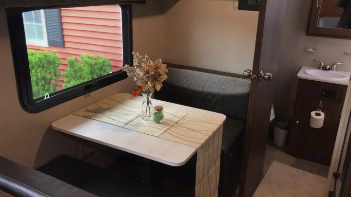 This rv rental features a cozy dinette with a window showing a house and bushes, adjacent to a compact bathroom.. Forest River Rockwood 2018