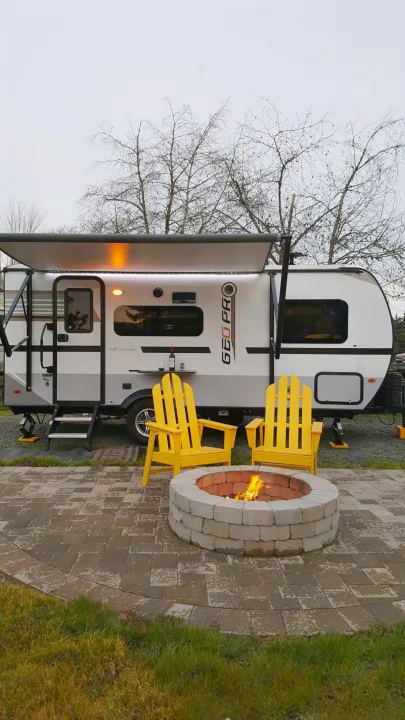 RV rental campsite with a white travel trailer, glowing fire pit, and two bright yellow chairs on a paved patio.. Forest River Rockwood Geo Pro 2018