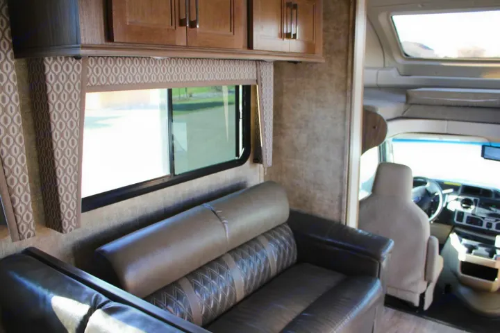 Interior of an RV rental: a cozy sofa, large window, overhead cabinets, and the bright driver's area are visible.. Jayco Envoy 2018