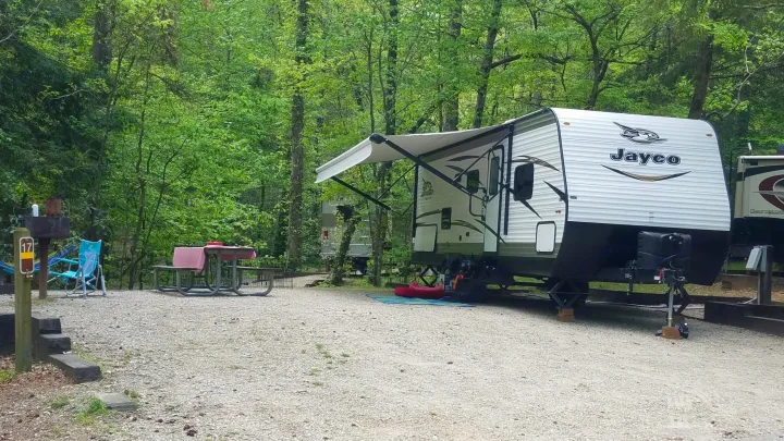 RV rental set up at a wooded campsite. A white Jayco travel trailer with an extended awning is parked on gravel, with a picnic area and chairs.. Jayco Jay Flight SLX 2018