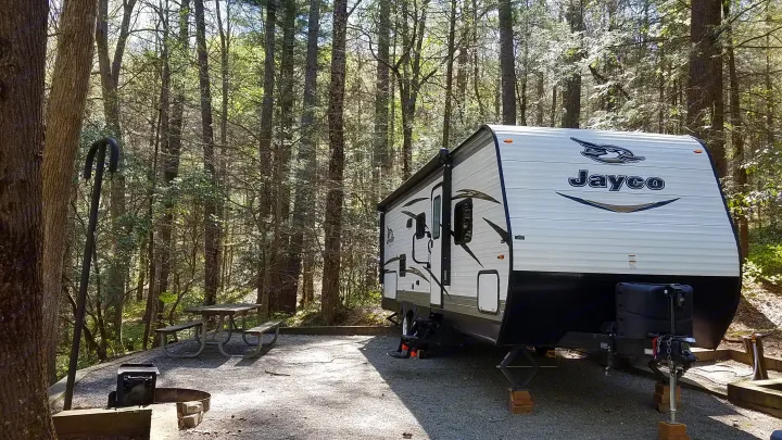 Jayco RV rental parked at a peaceful wooded campsite, featuring a picnic table and fire pit, ideal for camping.. Jayco Jay Flight SLX 2018