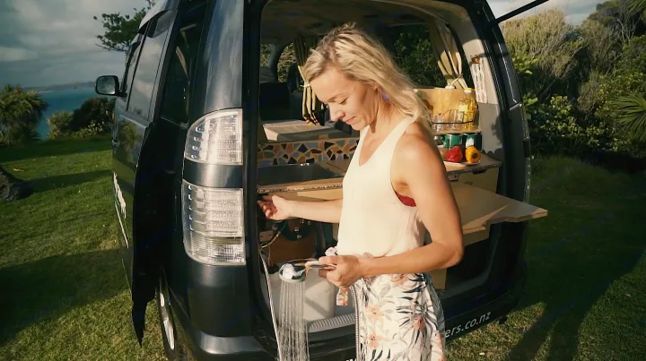 A woman uses her RV rental's outdoor shower and kitchen setup at the back of a camper van, parked on a grassy coastal hill.. Toyota Voxy 2006