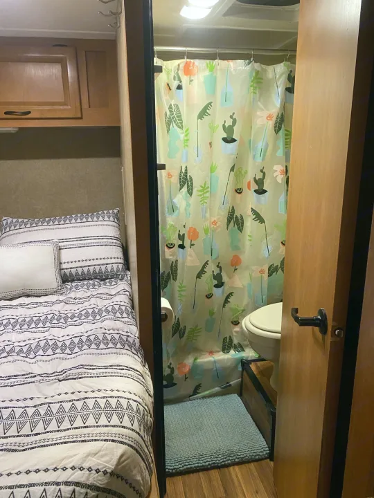 Compact RV rental interior featuring a cozy bed with patterned bedding and a brightly curtained bathroom with a toilet and bath mat.. Thor Motor Coach Four Winds Majestic 2015