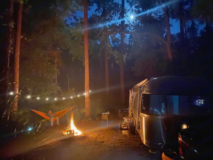 Cozy night camping scene with a person in a hammock by a campfire next to an RV rental. String lights & moonlight complete the ambiance.. Airstream International 2019