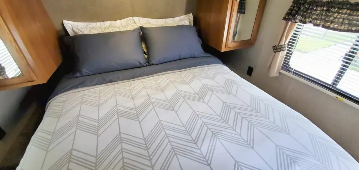 Cozy RV rental bedroom featuring a neatly made bed with gray chevron bedding, pillows, overhead cabinets, and a window.. Keystone Springdale 2017