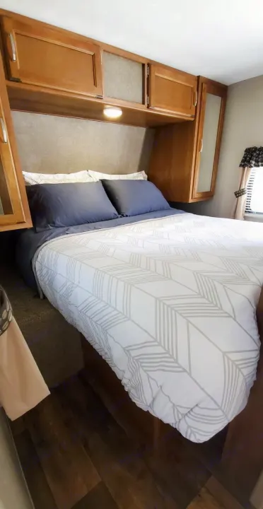 Comfortable RV rental bedroom featuring a bed with patterned comforter, dark pillows, and overhead wooden storage. Ready for your adventure!. Keystone Springdale 2017