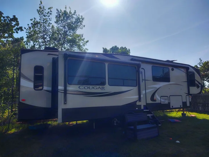Large beige Cougar fifth-wheel RV rental, parked on a sunny lawn with trees. Perfect for your outdoor escape.. Keystone Cougar 2019