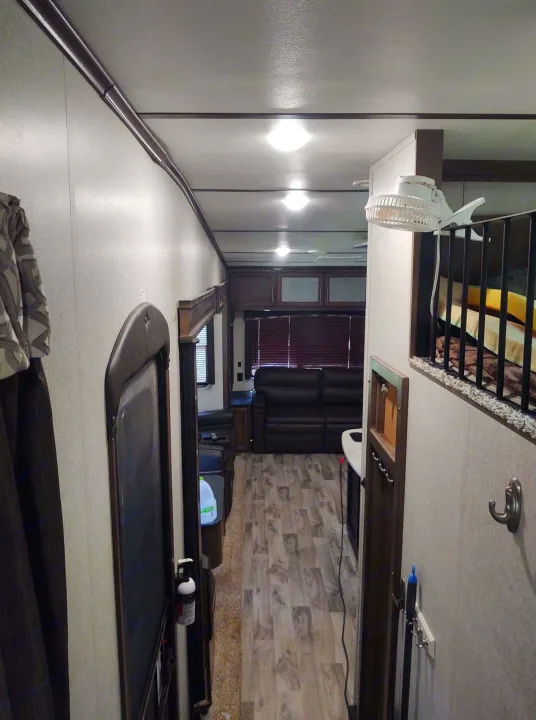 An RV rental's interior: a hallway view showing the living room with a couch, blinds, and an overhead bunk.. Keystone Cougar 2019