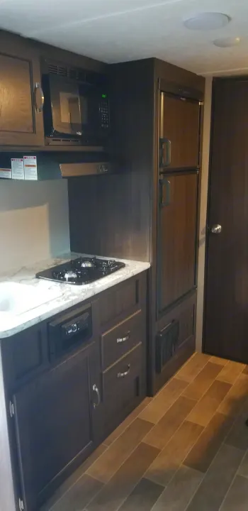 Inside this RV rental: a compact kitchen with dark wood cabinets, microwave, stovetop, and refrigerator.. Keystone Springdale 2019
