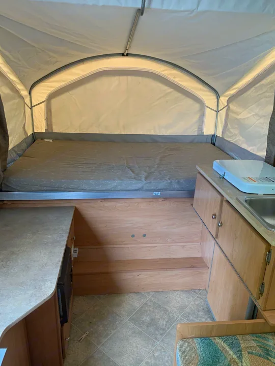 RV rental pop-up camper interior: extended bed, small kitchen with sink/stove, and wooden steps. Compact & functional.. Coachmen Sport 2007