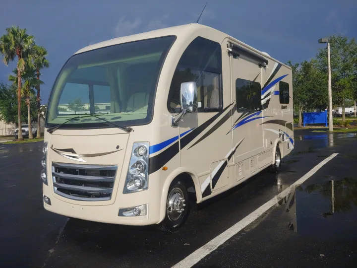 A beige Class C RV rental with bold blue and black graphics, parked on a wet lot after a recent rain. Perfect for trips!. Thor Motor Coach Vegas 24.1 2020