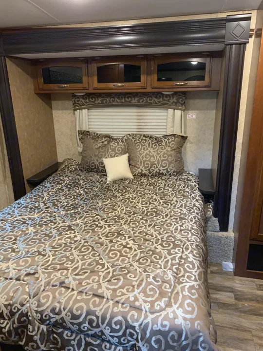 This RV rental bedroom features a comfy bed with intricate patterned bedding, overhead cabinets, and convenient side shelves.. Jayco White Hawk 2017