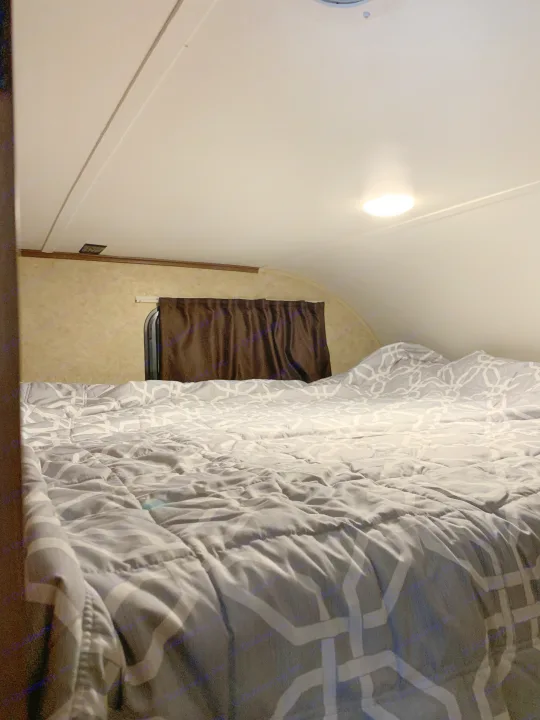 Cozy over-cab bed in an RV rental, featuring grey geometric bedding and window with brown curtains.. Jayco White Hawk 2017