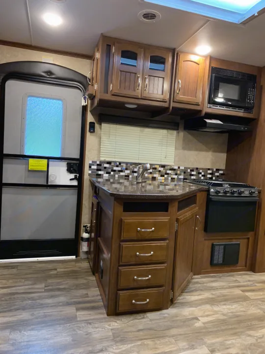 RV rental kitchen: Features a compact counter with sink, microwave, stove, and entrance door. Wood cabinets and flooring.. Jayco White Hawk 2017