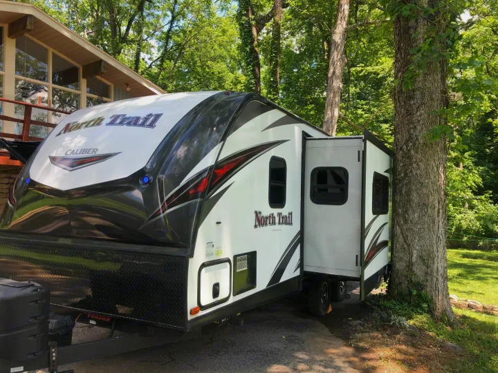 Beautiful North Trail RV rental, parked near a modern home amidst tall, green trees. Perfect outdoor escape.. Heartland North Trail 2019