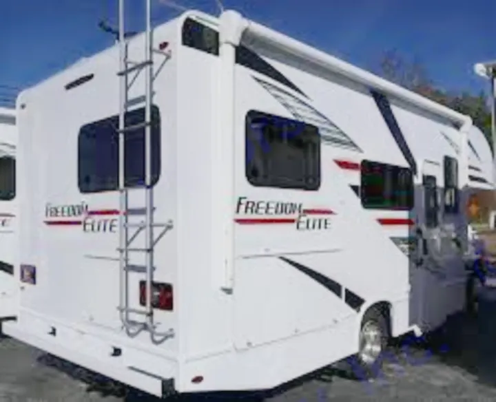 A white Freedom Elite Class C RV rental, viewed from the rear and side, features an extended awning and ladder.. Thor Freedom Elite 2020