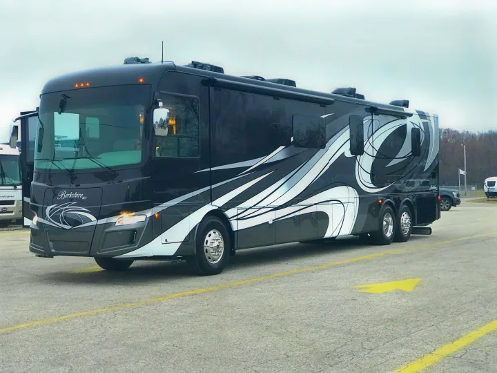 A sleek, black Berkshire XLT luxury motorhome, ideal for an RV rental, parked on asphalt. It features silver and white decals and multiple rear wheels.. Forest River Berkshire 2020