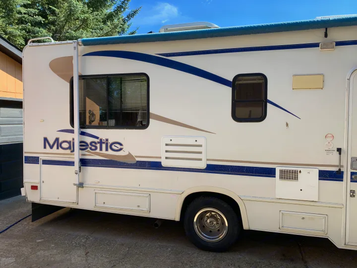Side view of a white Majestic RV rental with blue and tan stripes. Decals show extensive cracking, parked outdoors.. Majestic Leisure Craft E350 2001