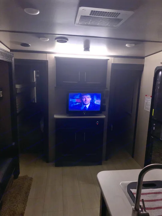 An rv rental's cozy interior: dim lighting highlights a TV, dark cabinets, hallway, and kitchen sink area for travel.. Forest River X-Lite 2018
