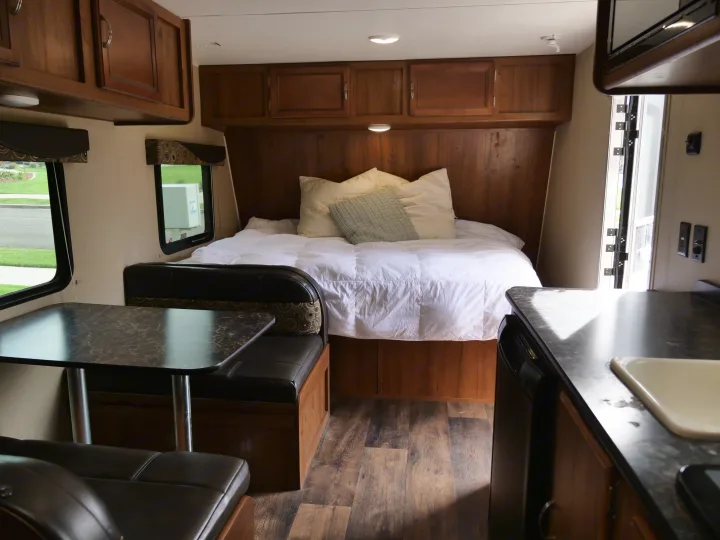 Inside an RV rental, showing a bed with wood cabinetry, a dinette with dark seating, and a kitchen counter with a sink.. Gulf Stream Innsbruck 2016
