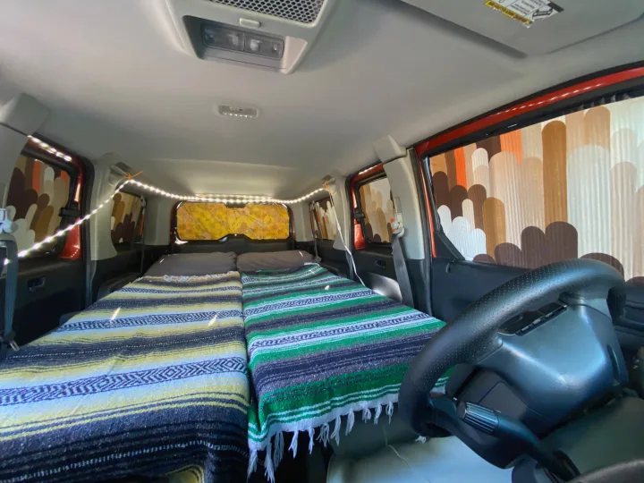 Interior of an RV rental with two cozy beds, colorful striped blankets, string lights, and patterned window covers, ideal for car camping.. Honda Element 2005