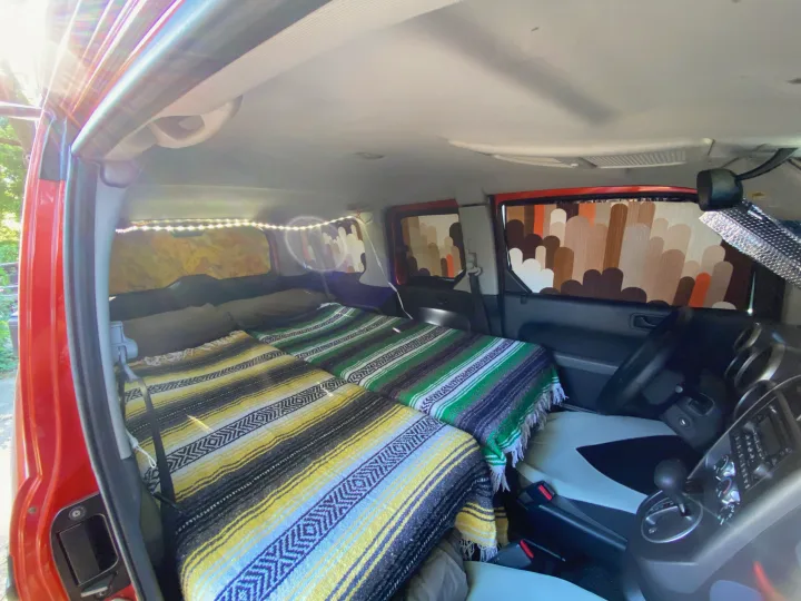 Inside a modified red vehicle, featuring a cozy camper bed setup with colorful blankets and LED lighting, ideal for an RV rental.. Honda Element 2005