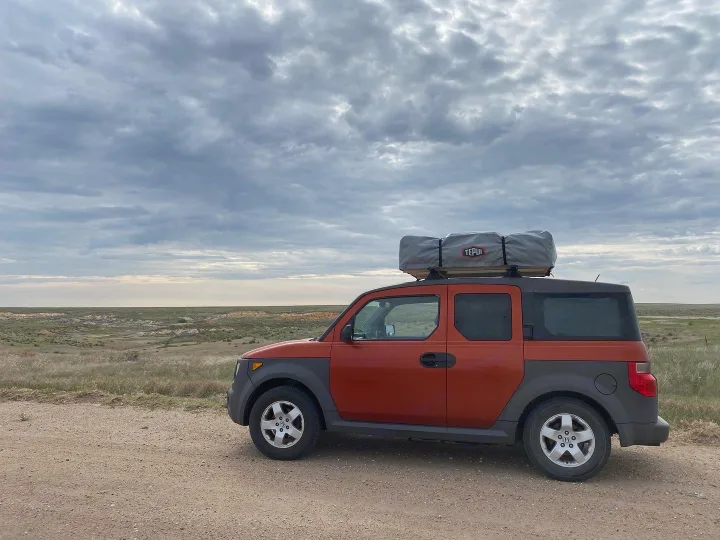 Red Honda Element with rooftop tent on a dirt road, vast plains under cloudy skies. Great for adventure travel, an RV rental alternative.. Honda Element 2005