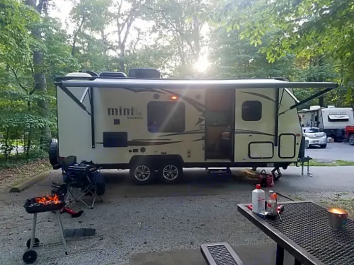 RV rental parked at a wooded campsite. A travel trailer with an extended awning, a lit grill, and picnic table are visible.. Forest River Rockwood Mini Lite 2017