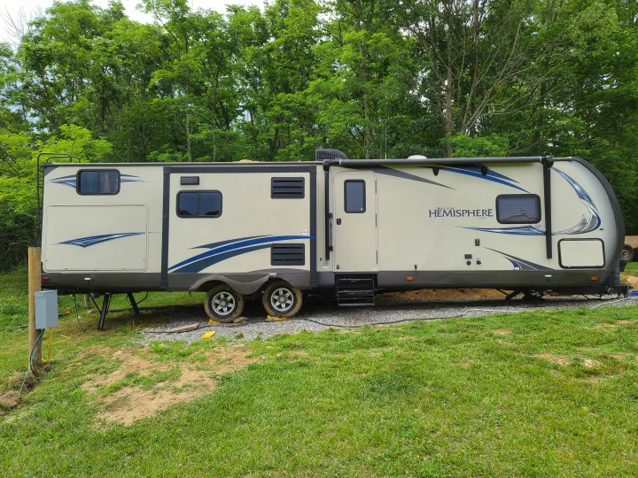 This beige travel trailer, ideal for an RV rental, is parked on gravel in a natural setting with lush green trees.. Forest River Salem Hemisphere 2015