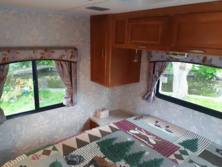 Cozy RV rental bedroom with a nature-themed quilt, wooden cabinets, and windows overlooking a pond.. Other Other 2009